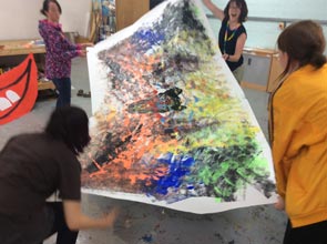 Four people open out a huge sheet of white paper covered in colourful paint