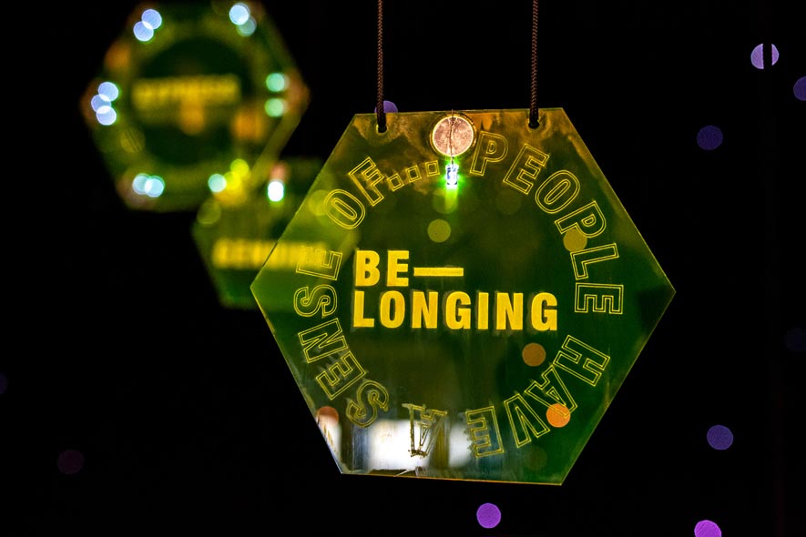 A suspended illuminated yellow hexagon etched with the words ‘People have a sense of belonging’ and suspended in a dark space with starlight in the background