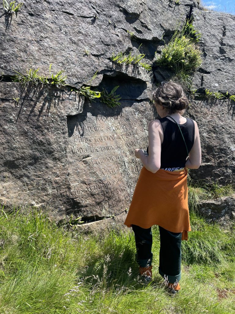Lucy standing in front a flat vertical rock (one of the Brontë Stones) with writing carved into it.
